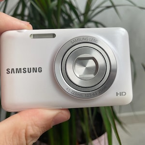 Puede incluir: Una cámara digital Samsung blanca con un objetivo plateado que lleva impreso el texto Samsung Lens, 4.5-22.5mm, 1:2.5-6.3, 25mm y HD. El cuerpo de la cámara es liso y rectangular, con un flash integrado situado sobre el objetivo.