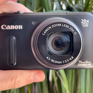 May include: A black Canon PowerShot SX260 HS digital camera featuring a 20x optical zoom lens. The front of the camera displays the text Canon, 20x Optical Zoom, Canon Zoom Lens 20x IS, 4.5-90.0mm 1:3.5-6.8, and PowerShot SX260 HS.