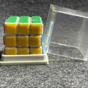 Op de afbeelding: Een kleurrijke Rubik's Cube met groene, gele, oranje en blauwe vierkanten, gepresenteerd op een witte basis. Een doorzichtige plastic doos staat naast de kubus. De puzzel is een klassieke hersenkraker.