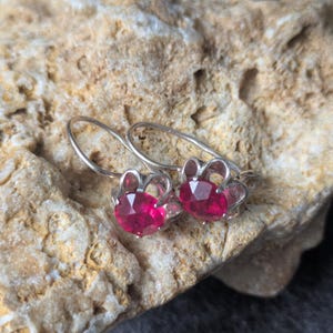 May include: A pair of silver earrings with a flower design. Each earring features a vibrant, round, ruby-red gemstone set within a delicate, openwork silver frame. The earrings are displayed on a textured, light-brown stone surface.