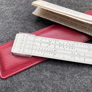 May include: A vintage slide rule with a white scale and clear plastic cursor, on a red leather case. A paper instruction manual is on top. The slide rule features numerical markings and scales for calculations.