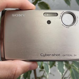 May include: A silver Sony Cyber-shot digital camera with a Carl Zeiss Vario-Tessar lens, featuring an optical 3x zoom, a built-in flash, and a small circular sensor on the front face, with the brand name and model text clearly printed on the body.