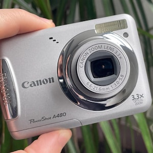 May include: Silver Canon PowerShot A480 digital camera. Features a Canon Zoom Lens 3.3x with a focal length of 6.6-21.6mm. Includes a built-in flash and a 3.3x optical zoom. The camera is held in a hand.