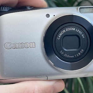 May include: Silver Canon digital camera with a black lens and a built-in flash. The camera has a 4x optical zoom and 14.1 megapixels. The Canon logo is visible on the front.