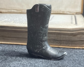 May include: A small, dark gray cowboy boot figurine with intricate floral and scrollwork detailing. The boot has a pointed toe and a small heel. The figurine is made of metal and appears to be vintage or antique.