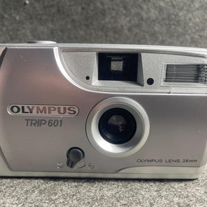 Op de afbeelding: Zilveren Olympus Trip 601 camera met een 28mm lens. De camera heeft een rechthoekige behuizing met een lens, flitser en zoeker. De merknaam en het modelnummer staan op de voorkant.