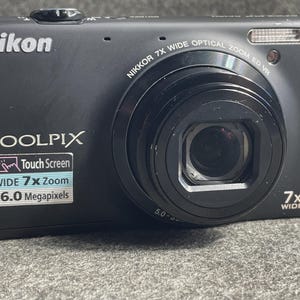 May include: Black Nikon Coolpix digital camera with a wide 7x zoom lens. Features a touch screen and 16.0 megapixels. The camera has the Nikon and Coolpix brand names on the front.