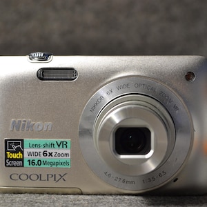 Könnte beinhalten: Silberne Nikon Coolpix Digitalkamera mit 6x Zoom und 16,0 Megapixeln. Mit Touchscreen und Lens-Shift VR. Das Objektiv hat 4,6-27,6 mm. Rechts daneben eine kleine Spielfigur.
