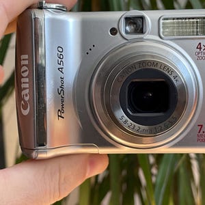 Op de afbeelding: Een zilverkleurige Canon PowerShot A560 digitale camera met een 4x optische zoomlens, 7,1 megapixels en een 5,8-23,2 mm 1:2,6-5,5 lens. De voorzijde van de camera toont de merknaam, het modelnummer en technische specificaties in zwarte tekst.