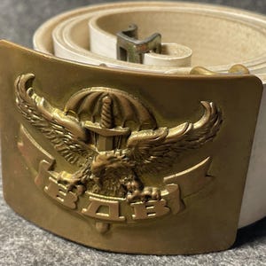 May include: A white belt with a brass buckle. The buckle has an eagle design with a sword and the letters "ВДВ". The buckle is rectangular with a curved top and bottom.