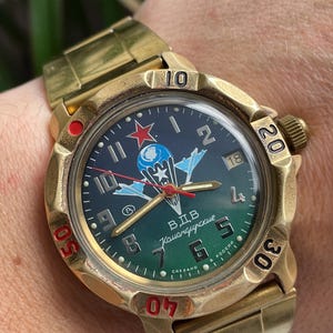 Puede incluir: Un reloj de pulsera dorado con una esfera verde oscuro y azul. La esfera del reloj presenta una estrella roja, un emblema de paracaídas y el texto "В.Д.В. Камандирские". El reloj tiene una correa dorada y un bisel giratorio.