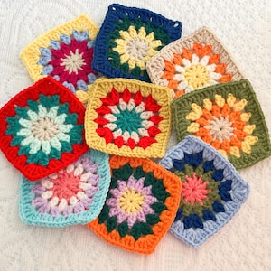 May include: A collection of colorful, square crochet pieces in various shades, including red, blue, yellow, and green. Each piece features a central floral design with contrasting borders. The handmade items are arranged on a white, textured surface.