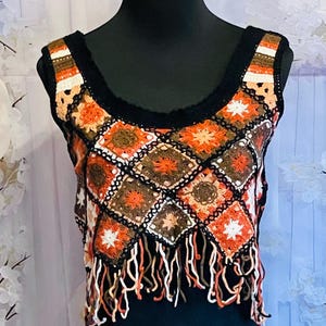May include: A sleeveless crochet top featuring a pattern of square motifs in shades of orange, brown, white, and black. The garment has a black neckline and a hemline finished with long, dangling yarn fringe in matching colors.