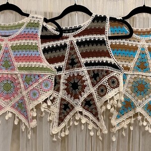 May include: Three handmade crochet vests in various colors, including pink, green, brown, black, and blue. Each vest features a star design with a fringed hem and is displayed on a black hanger.