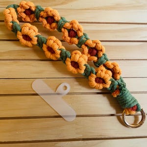 May include: A handmade macrame phone strap featuring a repeating pattern of orange and burgundy flower-shaped knots with green leaves. The strap has a gold-colored metal ring and a clear plastic phone holder.