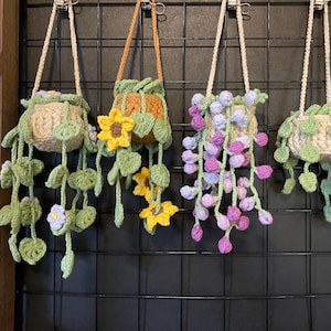 May include: Four handmade crochet hanging planters. Each planter is a different color and design, with cream, orange, and green pots. The planters are decorated with crochet flowers and vines, including sunflowers and purple flowers. The planters hang from a black grid.