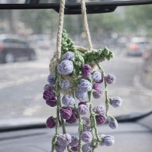 May include: A handmade crochet car charm featuring a small beige basket with green leaves and hanging purple and lavender flower clusters. The charm hangs from a beige rope and is suspended in front of a car's rearview mirror.