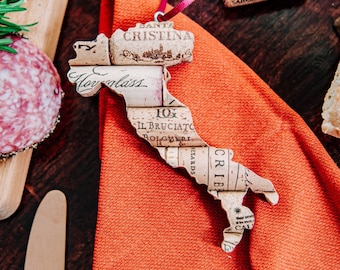 May include: A cork map of Italy in the shape of the country. The map is made of wine corks and has the names of Italian regions and cities written on it. The map is on a red fabric surface.