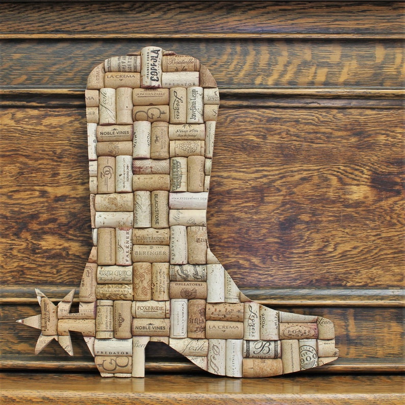 May include: A cowboy boot shaped wall art made from wine corks. The boot is brown and has a spur on the heel. The corks are arranged in a pattern and have various labels and text on them. Some of the text includes "La Crema", "Noble Vines", "Blackstone", "Beringer", "Foxbrode", "Stella", "Dellatore", "Ponzi", and "100%.