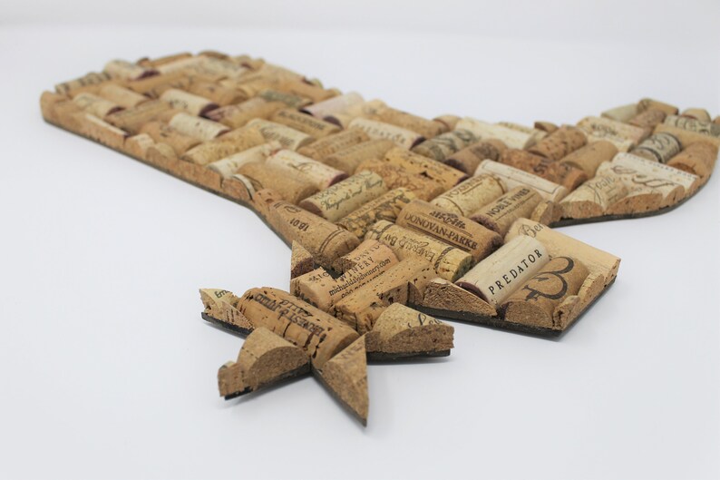May include: A cork art piece shaped like the state of Texas with a star in the bottom left corner. The cork pieces are arranged in a mosaic pattern and have various wine labels and text on them. The text on the cork pieces includes "PREDATOR", "J.J.B.R.  MONDOVAN-PARKE", and "WORLD WINE".