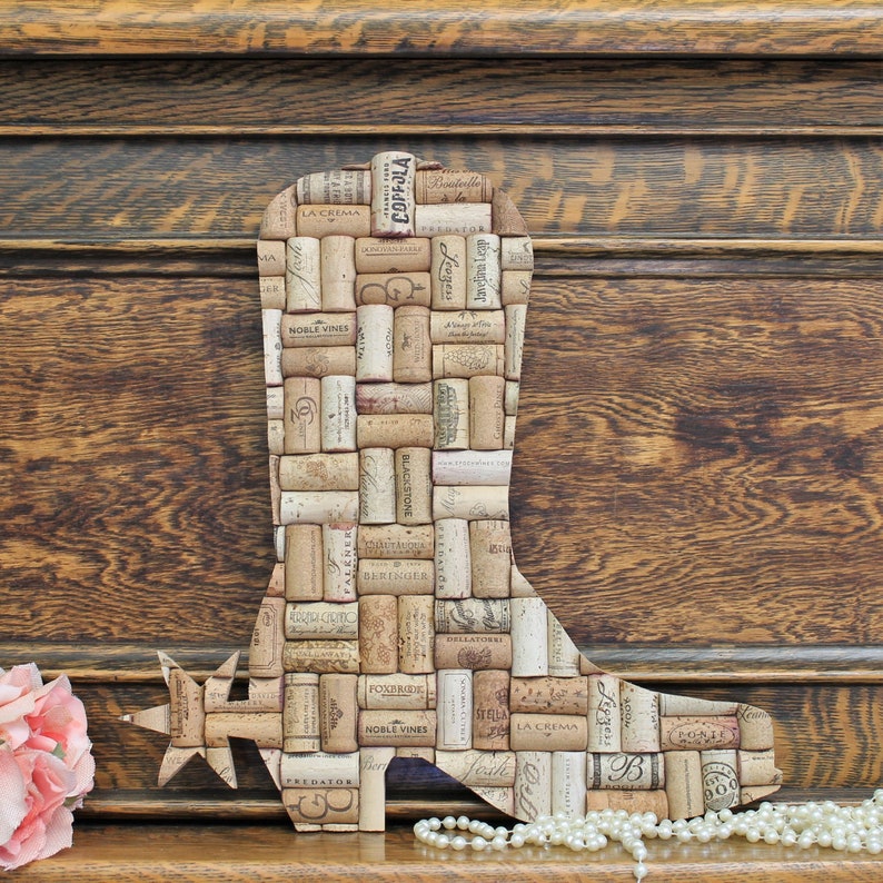 May include: A cowboy boot shaped wall decor made from wine corks. The boot is brown and has a spur on the heel. The corks are arranged in a pattern and have various labels and text on them.