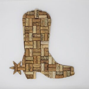 May include: A cowboy boot shaped wall art made from wine corks. The boot has a spur on the heel and is decorated with various wine cork labels.