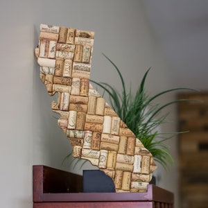 May include: A cork map of the state of California, made from wine corks. The map is mounted on a wooden base and is decorated with various wine labels. The map is displayed on a wooden shelf.