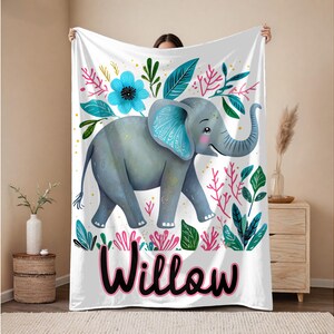 Peut inclure: Une couverture de b&eacute;b&eacute; blanche avec une illustration color&eacute;e d'un &eacute;l&eacute;phant aux oreilles bleues et une fleur rose. La couverture porte le nom "Willow" imprim&eacute; en noir en bas.
