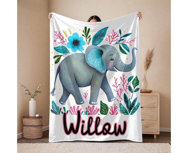 Peut inclure: Une couverture de b&eacute;b&eacute; blanche avec une illustration color&eacute;e d'un &eacute;l&eacute;phant aux oreilles bleues et une fleur rose. La couverture porte le nom "Willow" imprim&eacute; en noir en bas.