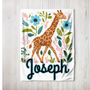 Peut inclure: Une couverture de b&eacute;b&eacute; blanche avec un motif de girafe et de fleurs. La girafe est brune et jaune avec un long cou et des taches. Les fleurs sont bleues, roses et vertes. Le nom "Joseph" est &eacute;crit en noir avec un contour bleu.