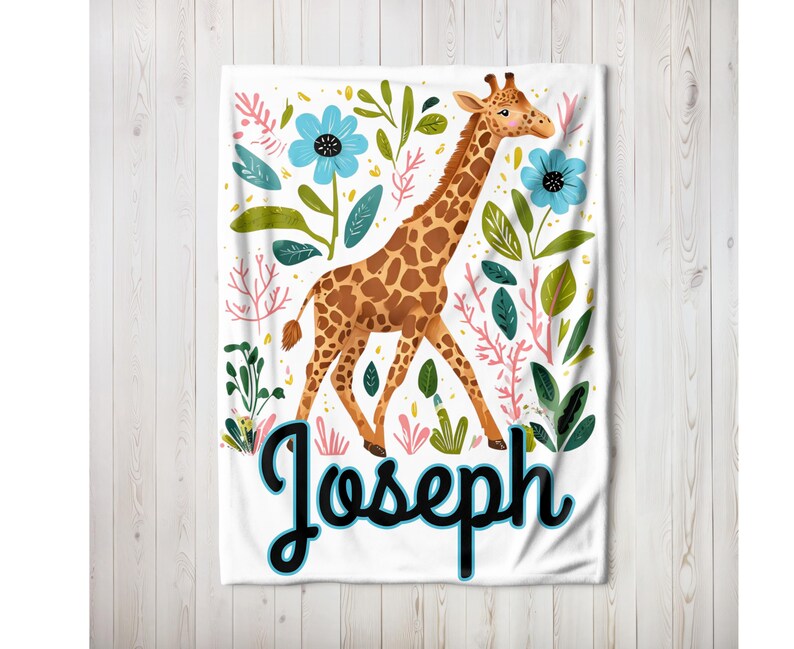 Peut inclure: Une couverture de b&eacute;b&eacute; blanche avec un motif de girafe et de fleurs. La girafe est brune et jaune avec un long cou et des taches. Les fleurs sont bleues, roses et vertes. Le nom "Joseph" est &eacute;crit en noir avec un contour bleu.
