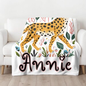 Peut inclure: Une couverture en polaire blanche avec un motif l&eacute;opard et des accents floraux. La couverture porte le nom "Annie" en &eacute;criture cursive noire.