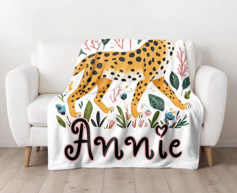 Peut inclure: Une couverture en polaire blanche avec un motif l&eacute;opard et des accents floraux. La couverture porte le nom "Annie" en &eacute;criture cursive noire.