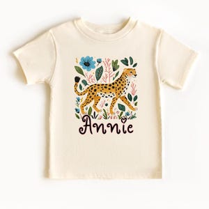 May include: A cream colored t-shirt with a cheetah print design and floral accents. The shirt has the name "Annie" printed on the front.