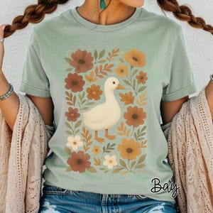 May include: Sage green t-shirt featuring a white duck illustration surrounded by brown and orange flowers and leaves. The duck has an orange beak and feet. The word "Bay" is written in cursive at the bottom right.
