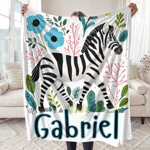 Peut inclure: Une couverture de b&eacute;b&eacute; blanche avec un z&egrave;bre noir et blanc, des fleurs roses et vertes et des fleurs bleues. La couverture porte le nom "Gabriel" imprim&eacute; en bas.