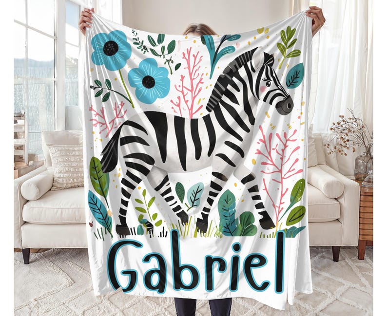 Peut inclure: Une couverture de b&eacute;b&eacute; blanche avec un z&egrave;bre noir et blanc, des fleurs roses et vertes et des fleurs bleues. La couverture porte le nom "Gabriel" imprim&eacute; en bas.