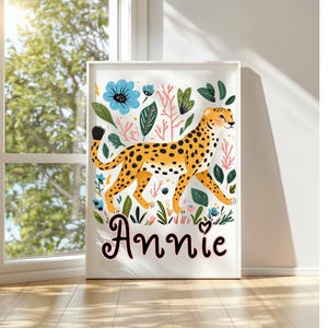Puede incluir: Un estampado art&iacute;stico enmarcado que presenta un guepardo estilizado con manchas naranjas, caminando entre flores y follaje colorido. El nombre "Annie" est&aacute; escrito en cursiva en la parte inferior. La obra tiene un borde blanco.