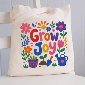 May include: A cream-colored canvas tote bag featuring a colorful graphic print with the text Grow Joy in large, bold letters. The design includes various illustrations of flowers, a potted plant, a small seedling, a garden trowel, and a watering can.
