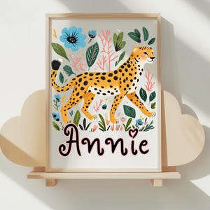 Puede incluir: Una impresi&oacute;n art&iacute;stica enmarcada que presenta un guepardo con pelaje amarillo y manchas negras, rodeado de flores azules y follaje verde. El nombre "Annie" est&aacute; escrito en una fuente decorativa. La obra de arte se exhibe en una estanter&iacute;a de madera.