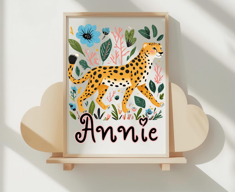 Puede incluir: Una impresi&oacute;n art&iacute;stica enmarcada que presenta un guepardo con pelaje amarillo y manchas negras, rodeado de flores azules y follaje verde. El nombre "Annie" est&aacute; escrito en una fuente decorativa. La obra de arte se exhibe en una estanter&iacute;a de madera.