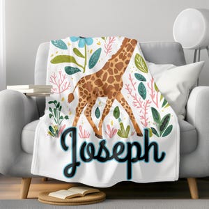 Peut inclure: Une couverture en polaire blanche avec une illustration color&eacute;e de girafe et des motifs floraux verts, roses et bleus. Le nom "Joseph" est &eacute;crit avec un contour bleu.