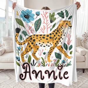 Peut inclure: Une couverture en polaire blanche avec une illustration color&eacute;e d'un gu&eacute;pard marchant dans une jungle avec des fleurs et des feuilles. La couverture porte le nom "Annie" imprim&eacute; en noir en bas.