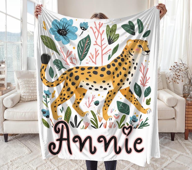 Peut inclure: Une couverture en polaire blanche avec une illustration color&eacute;e d'un gu&eacute;pard marchant dans une jungle avec des fleurs et des feuilles. La couverture porte le nom "Annie" imprim&eacute; en noir en bas.