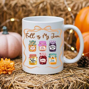 May include: White ceramic mug with the phrase "Fall is My Jam" and illustrations of various jam jars. The mug features images of peach jam, apple butter, blueberry jam, wildflower honey, preserves, and orange marmalade.