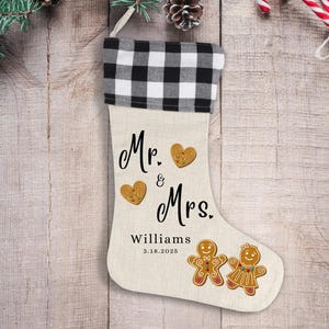 May include: A Christmas stocking with a black and white buffalo check cuff. The stocking is beige with the words "Mr. & Mrs. Williams 3.18.2025" and gingerbread people and heart accents.