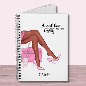 May include: A white spiral bound notebook with a pink and black illustration of a woman sitting with a pink handbag and a coffee cup. The text "A girl boss creating her legacy" and the name "Michelle" are printed on the cover.