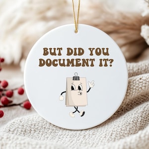 May include: A white ceramic ornament with the text "BUT DID YOU DOCUMENT IT?" in brown letters. The ornament features a cartoon notepad character with arms and legs. A gold string is attached for hanging.