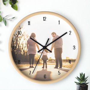 May include: Round wall clock with a light wood frame. The clock face features a family photo of a couple and a child walking on a path. Black clock hands point to the numbers around the clock face.