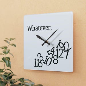 May include: Square white wall clock with black hands and the word "Whatever." printed in black. Black numbers are scattered across the clock face. The clock is mounted on a light orange wall.